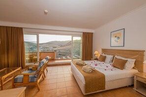 Habitación de hotel con balcón, cama con cubrecama blanco y marrón, cabecero de madera y un cuadro en la pared.