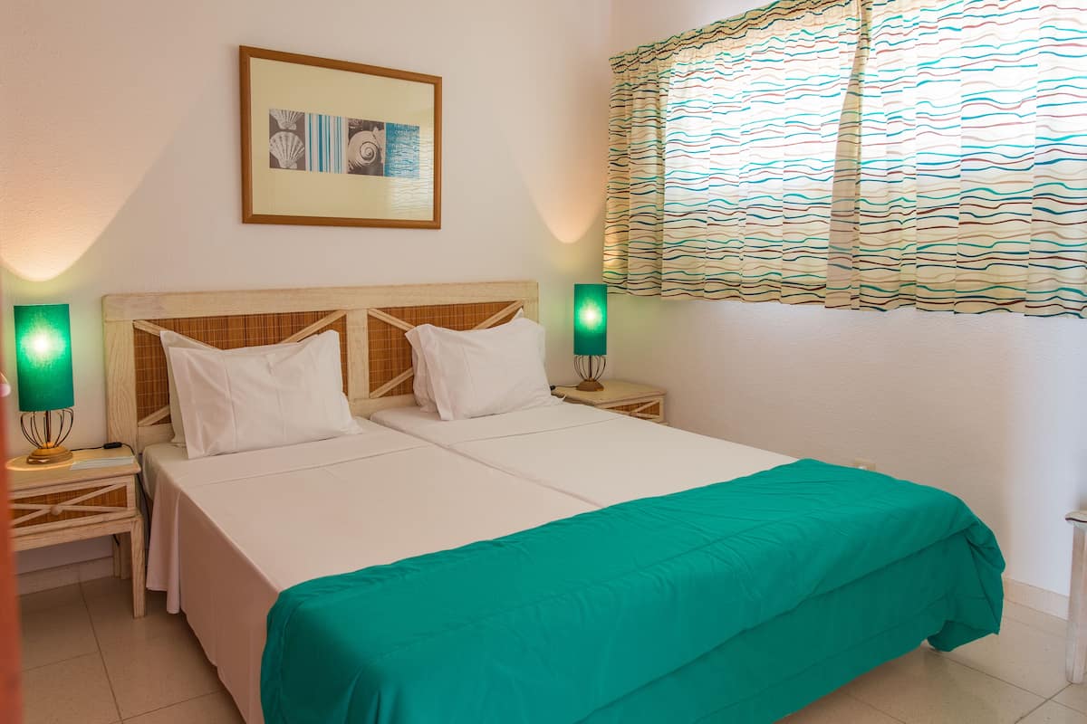 A neatly made bed with a green bedspread, two white pillows, and a wooden headboard. There are two bedside tables with lamps, and a framed picture on the wall above the bed.