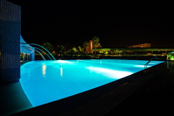 Una piscina nocturna con borde curvo, caída de agua y vista de un edificio de hotel al fondo.