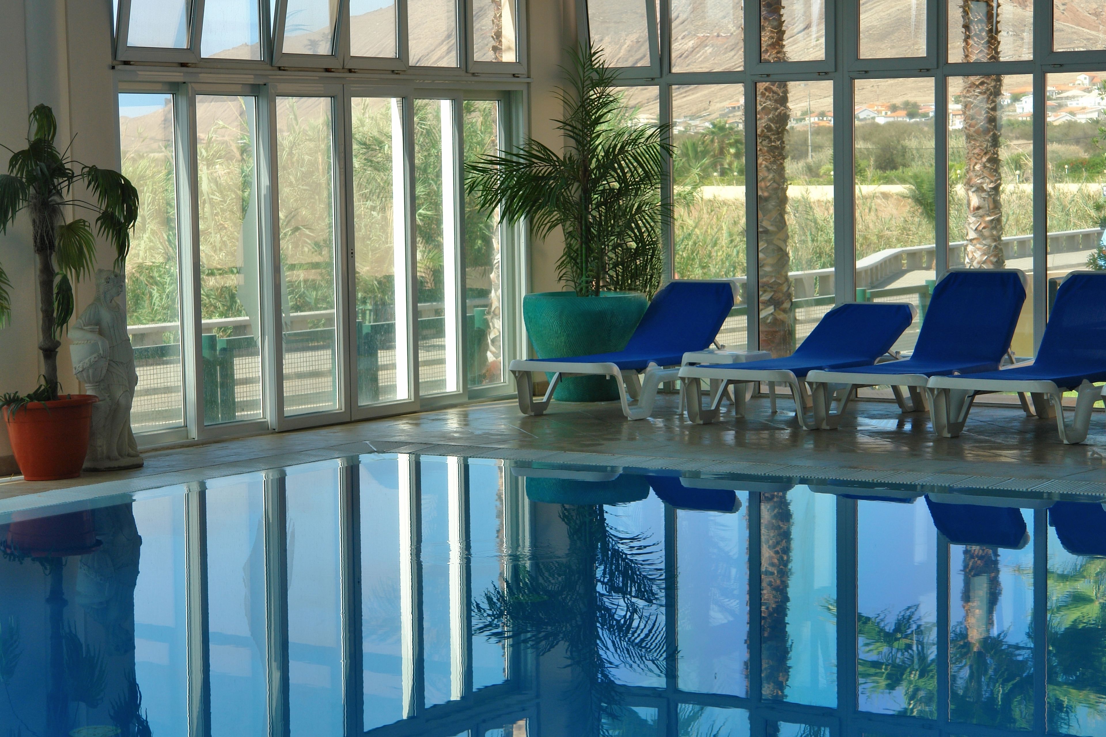 An indoor swimming pool with clear glass walls, blue lounge chairs, and potted plants.