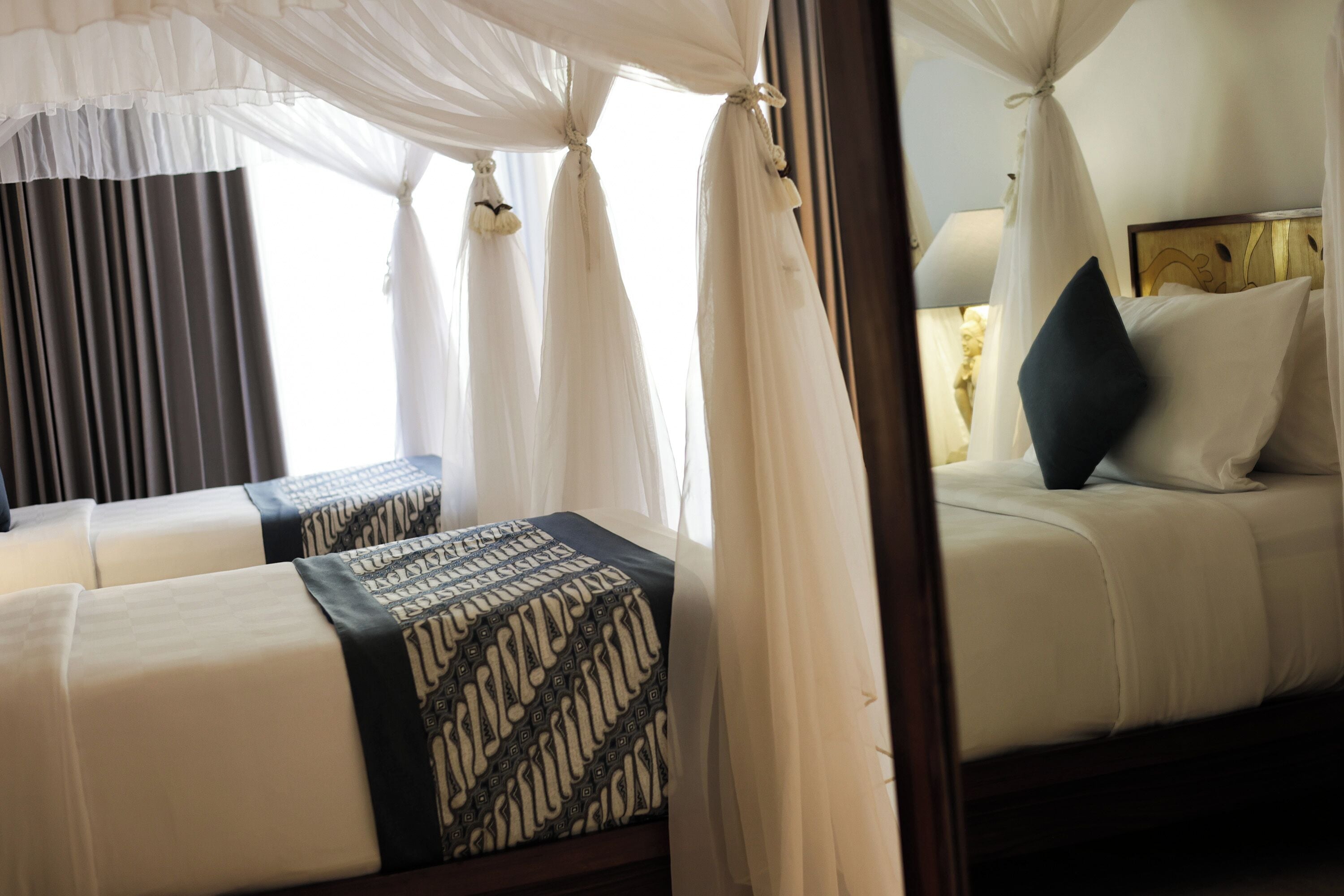 A hotel room with a canopy bed, two bedside tables with patterned cushions, and a view of the outside through sheer curtains.