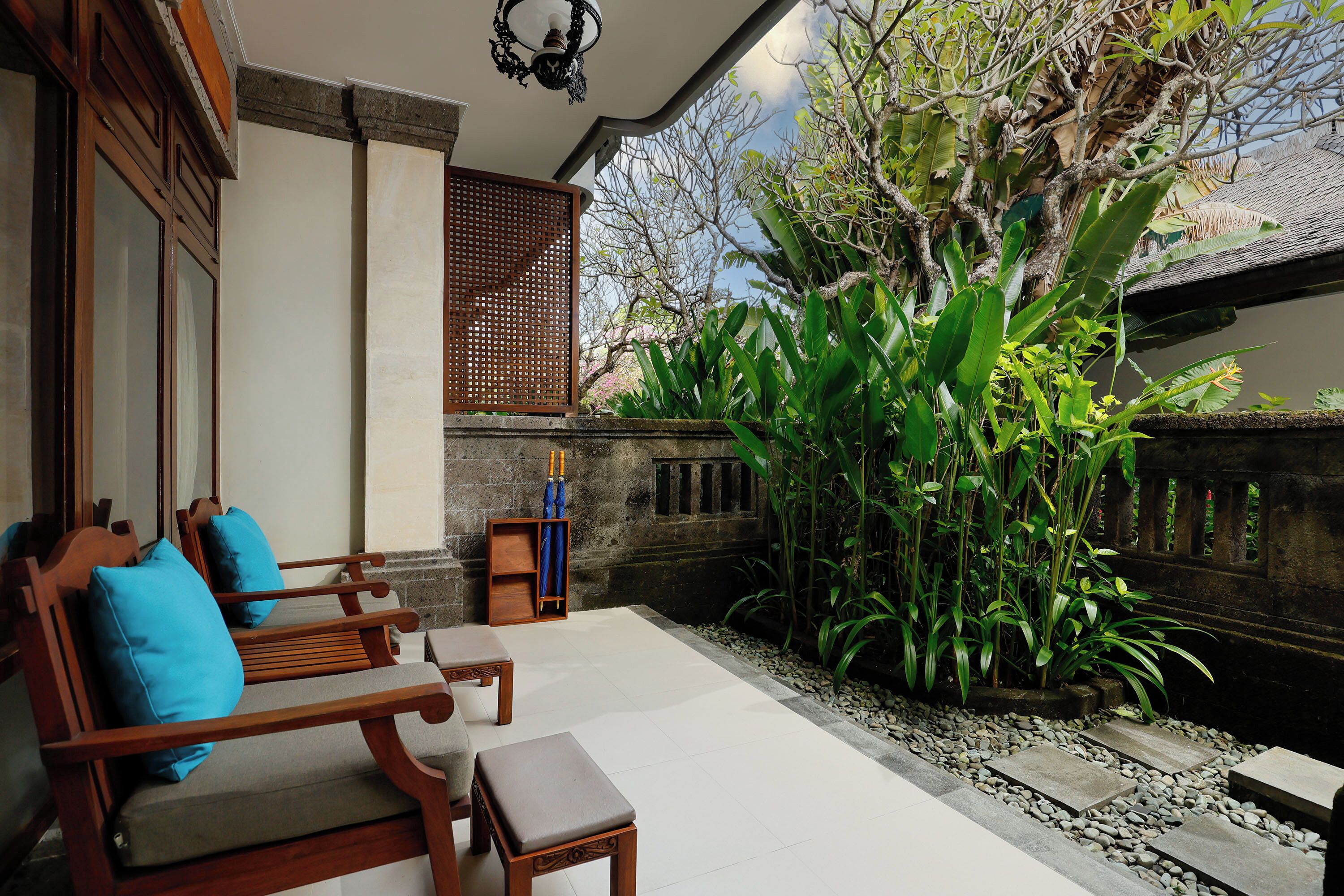 A wooden bench with cushions, a small table, and a potted plant in a courtyard with a stone pathway.