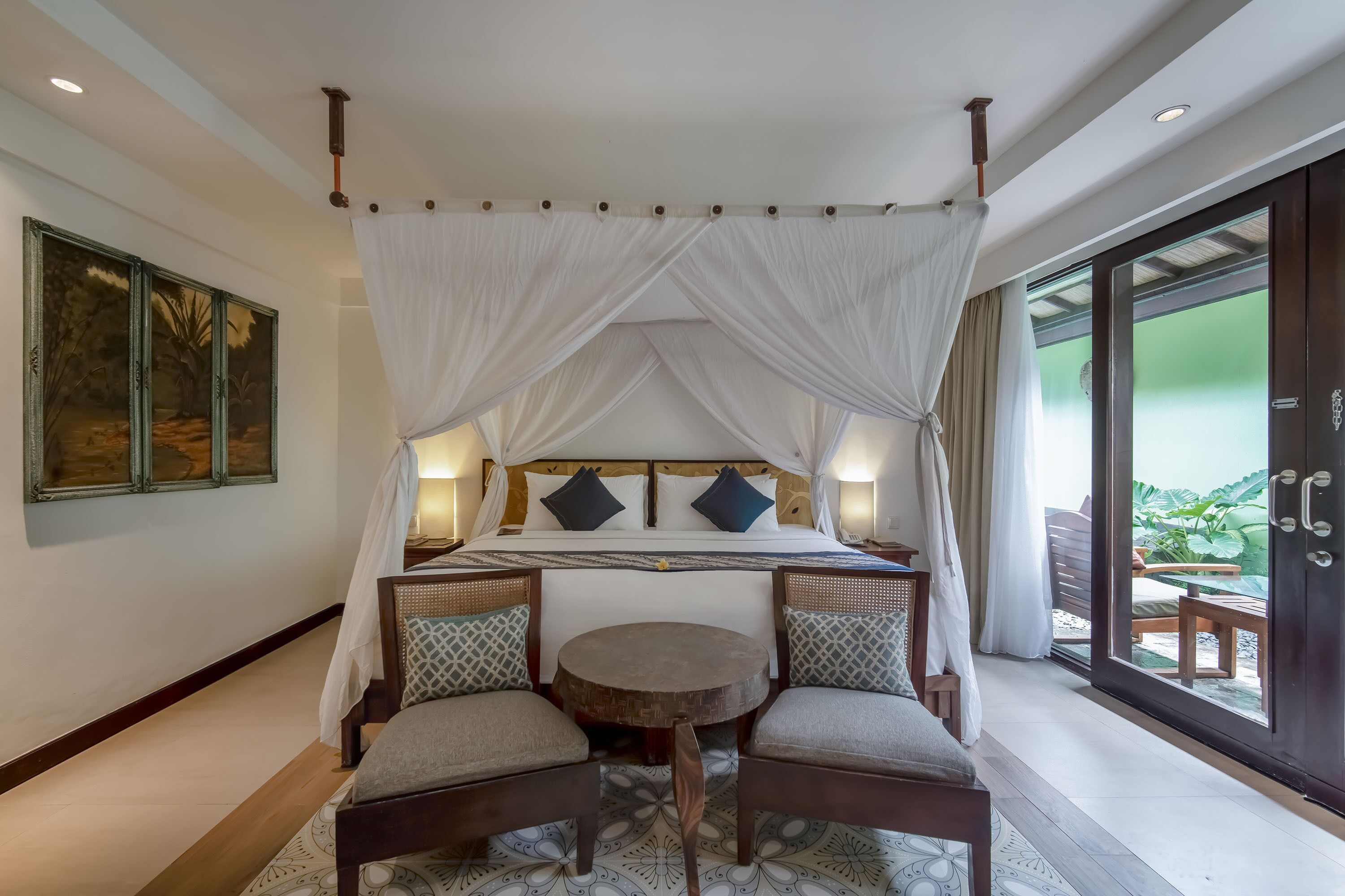 A room with a four-poster bed, a round table, two chairs, and a view through a glass door.