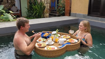 Um casal jantando em uma mesa flutuante na piscina, com diversos itens de café da manhã.