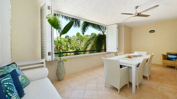 A room with a bed, a dining table with chairs, a ceiling fan, and a window with a view of greenery.