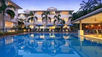 A swimming pool with lounge chairs and umbrellas, surrounded by a multi-story building with balconies and palm trees.