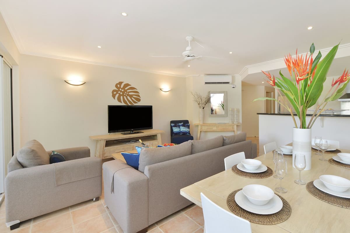 A modern living room with a flat-screen TV, a sofa, a dining table set for four, and a kitchen area in the background.