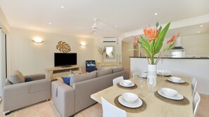 Uma sala de estar moderna com televisão de tela plana, um sofá, uma mesa de jantar para quatro pessoas e uma área de cozinha ao fundo.