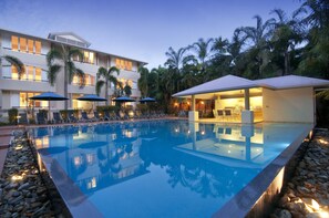 A poolside area with lounge chairs and umbrellas, a building with multiple balconies, and a clear pool reflecting the surroundings. - Cayman Villas (Port Douglas)