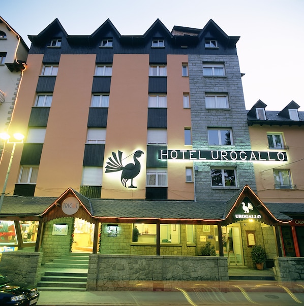 Un hotel llamado "Hotel Urogallo" con un logo de ave llamativo, fachada de piedra y una entrada tradicional con un letrero que dice "TRIC".