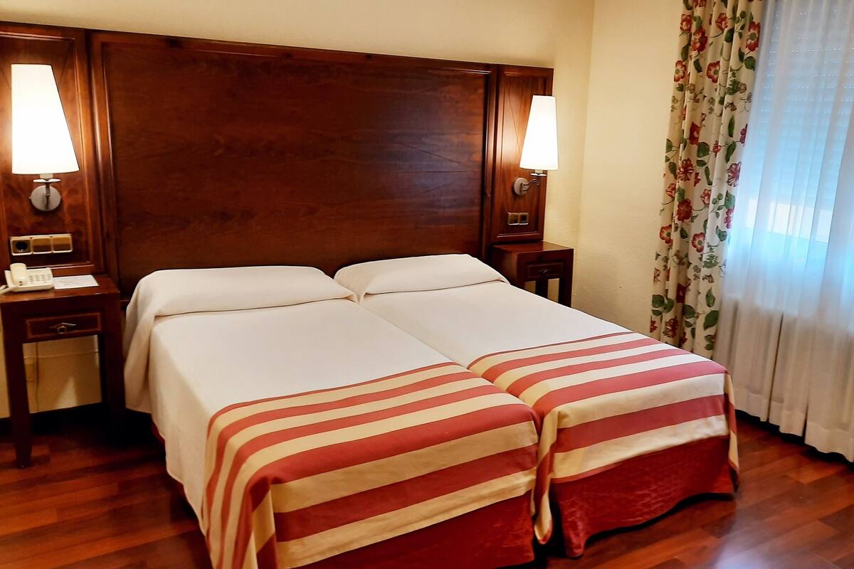 Een tweepersoonsbed met gestreepte lakens, een houten hoofdbord, nachtlampjes en bloemende gordijnen.