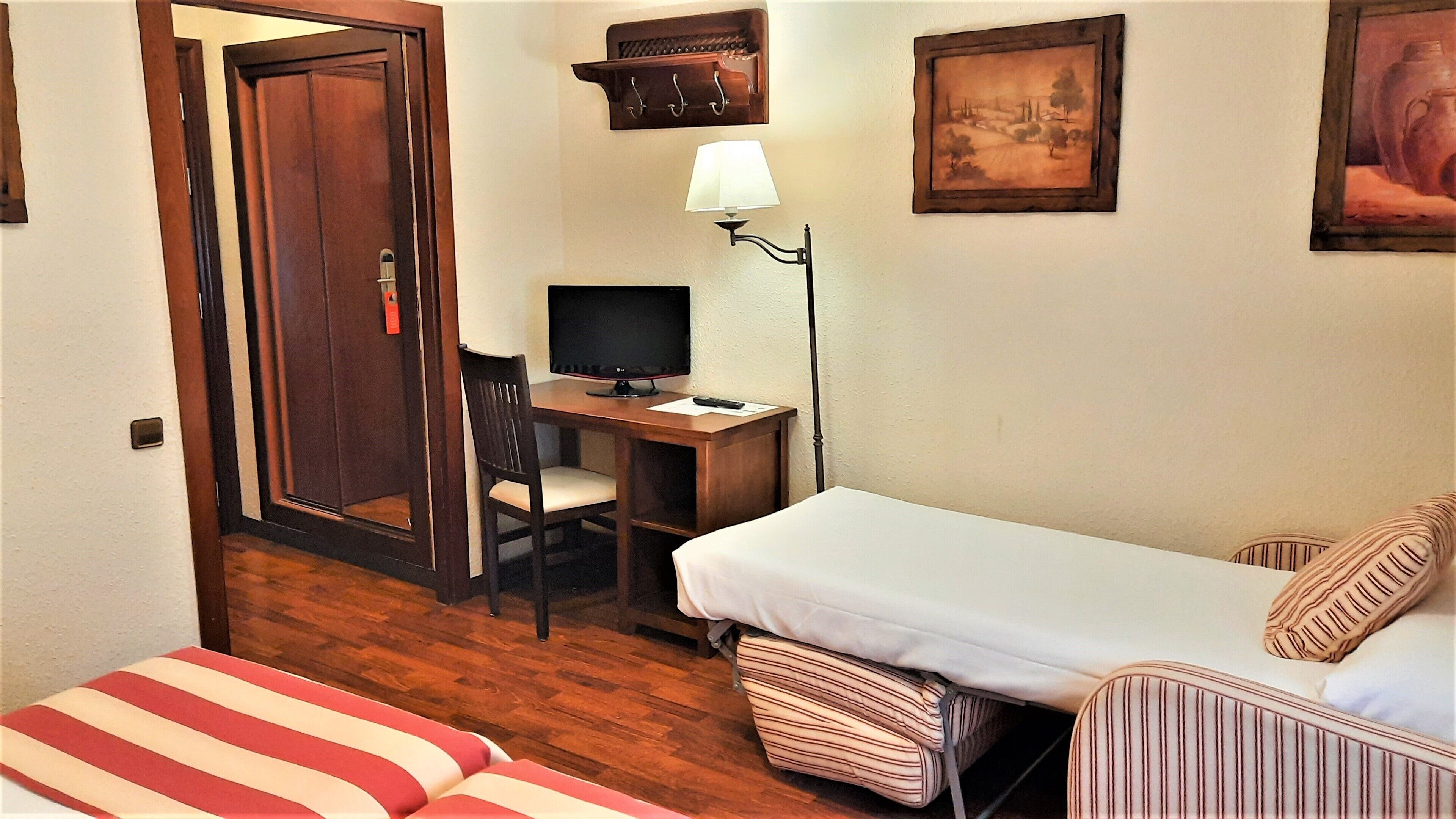A hotel room with a wooden floor, a bed with striped pillows, a desk with a computer and lamp, a chair, and a door with a red tag.