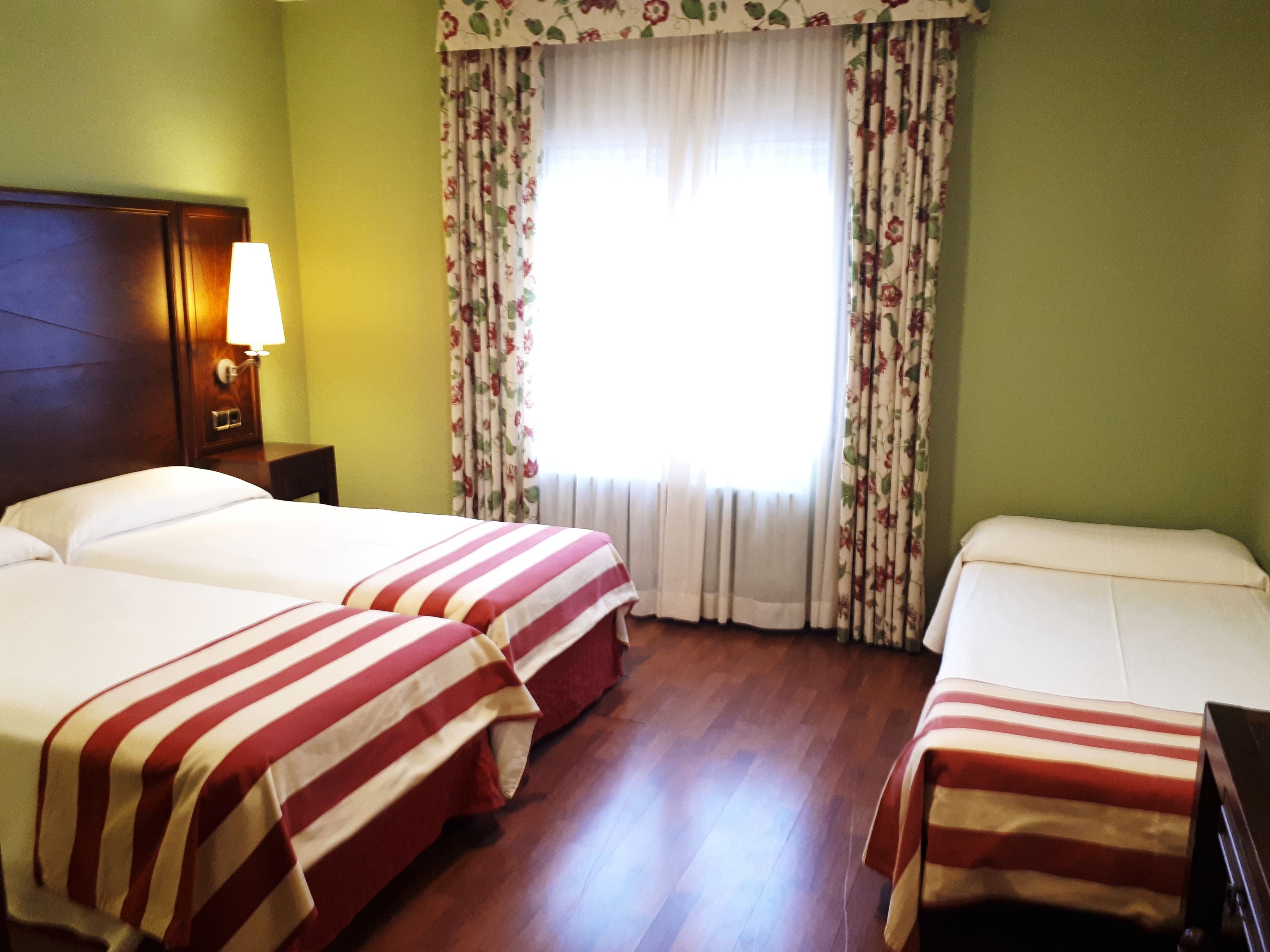 A hotel room with two beds, a nightstand, a lamp, and a window with floral curtains.