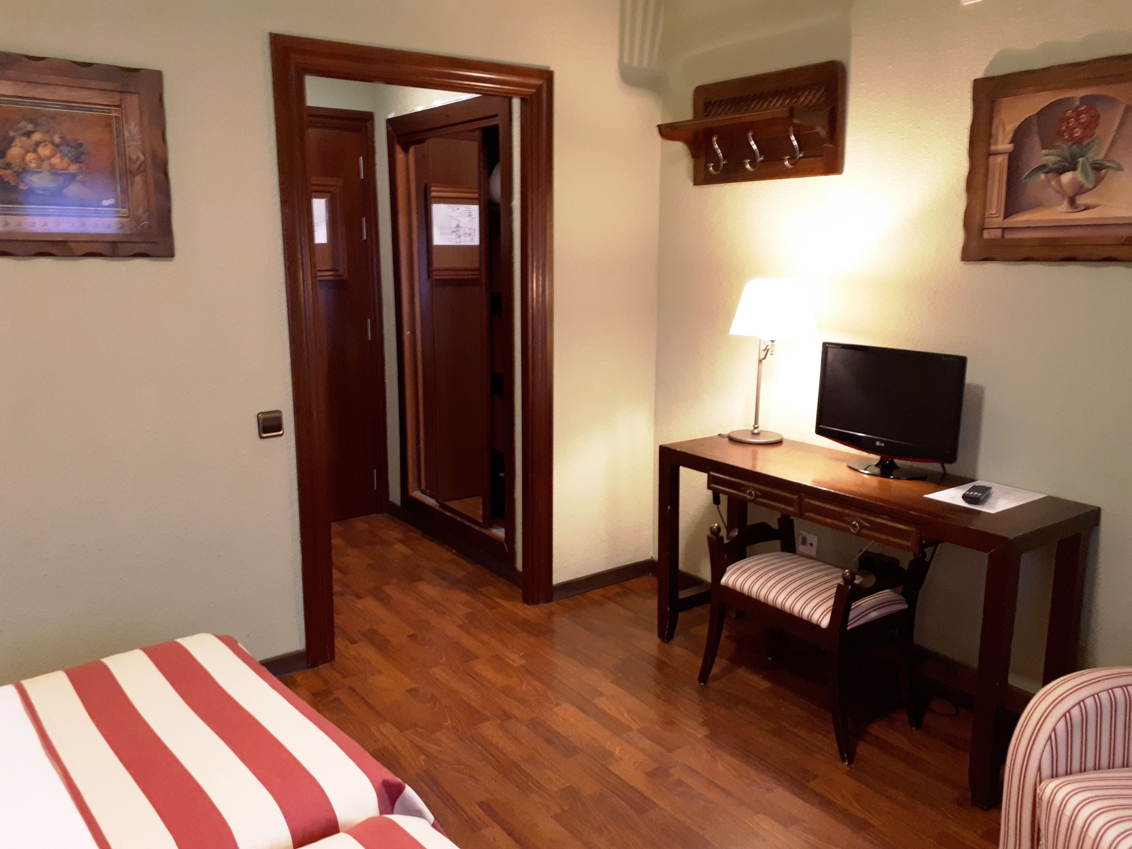 A hotel room with a wooden desk, a television, a striped chair, a bed, and a door leading to another room.