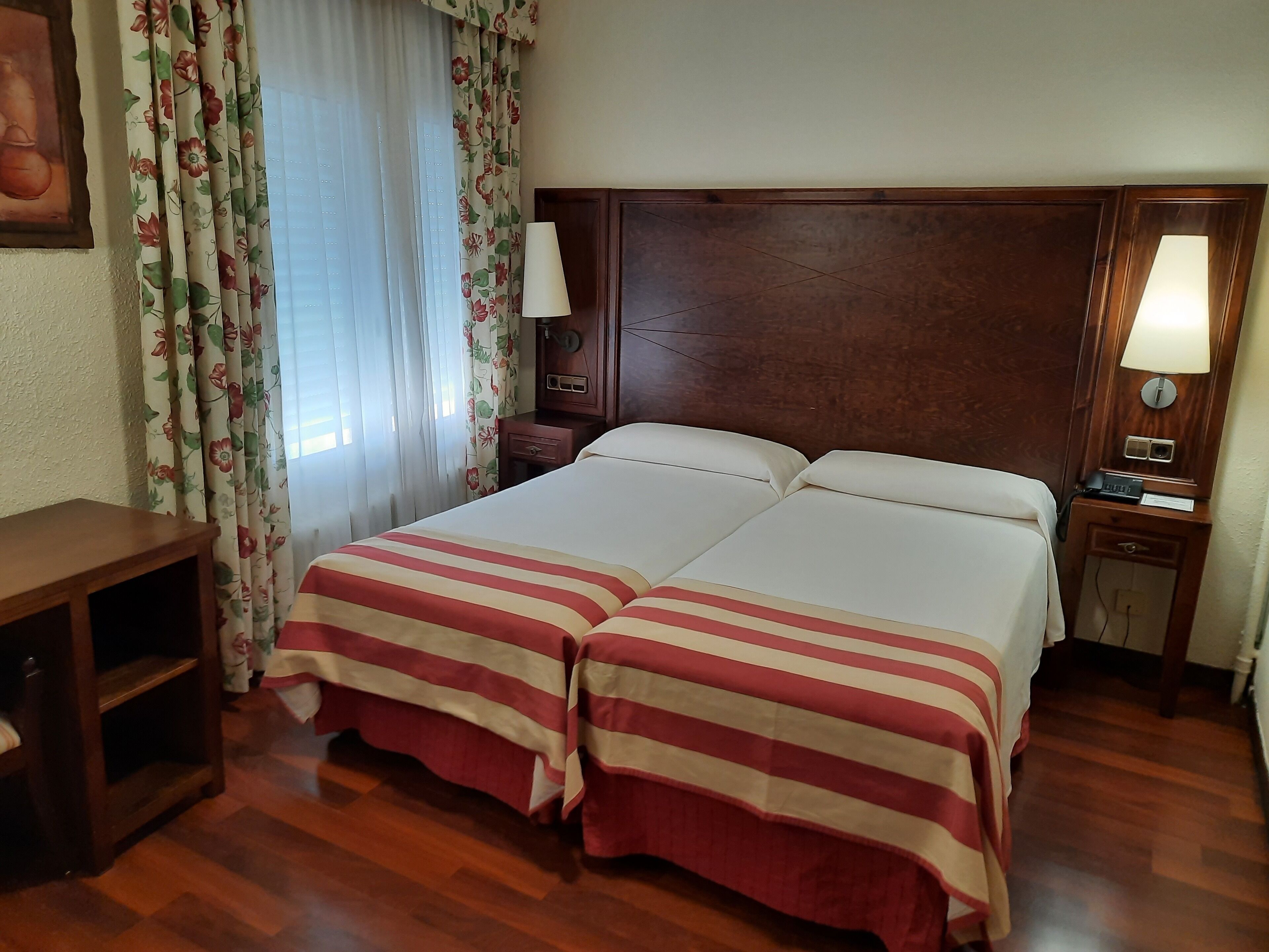Cama doble con ropa de cama a rayas, mesita de noche de madera con lámpara, escritorio con silla y cortinas con estampado floral.