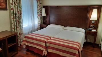 A double bed with striped bedding, a wooden nightstand with a lamp, a desk with a chair, and floral curtains.