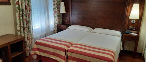 Cama doble con ropa de cama a rayas, mesita de noche de madera con lámpara, escritorio con silla y cortinas con estampado floral.