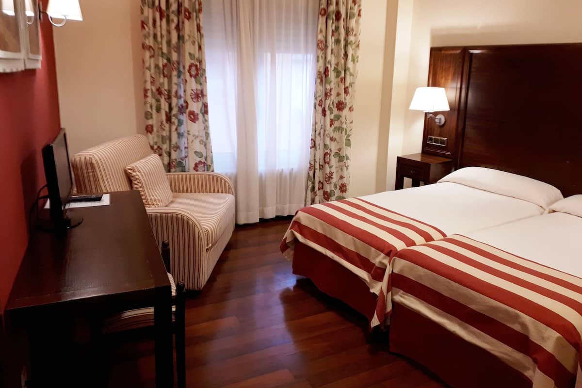 Een hotelkamer met een bed, een bureau met een computer, een stoel en een raam met bloemen gordijnen.