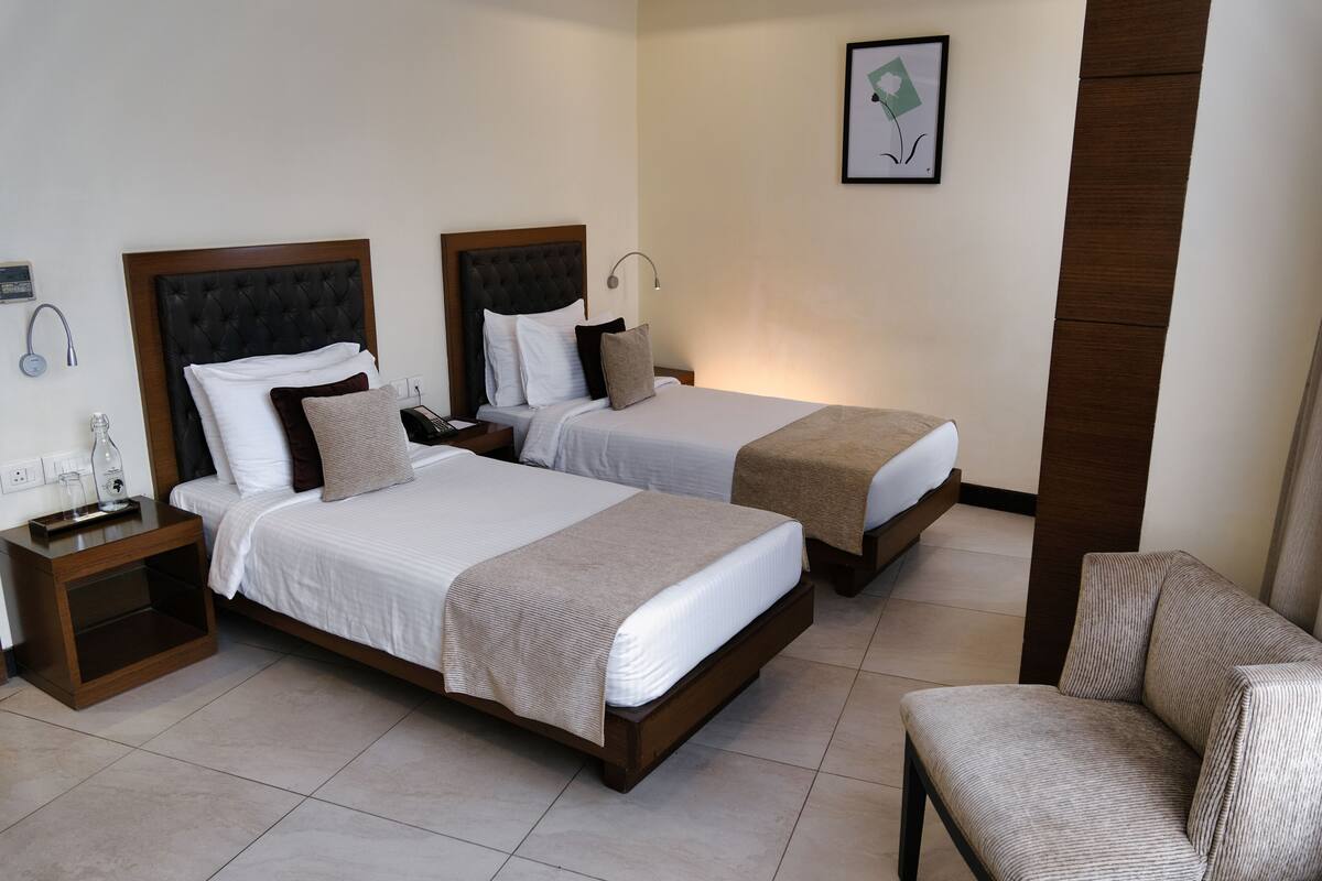Two single beds with wooden headboards, a bedside table with a lamp, a chair, and a framed picture on the wall.