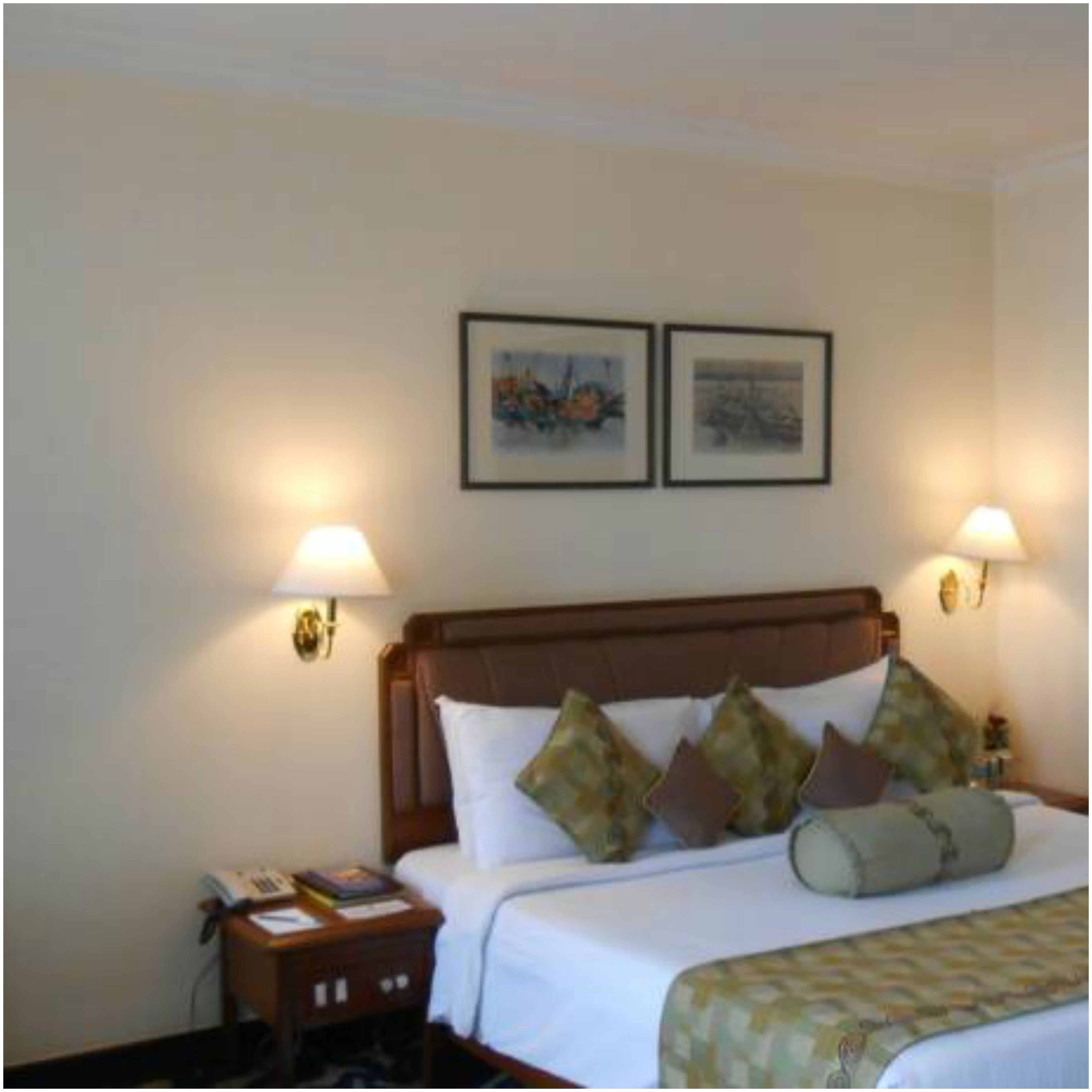 Une chambre d’hôtel avec une tête de lit en bois, un lit avec une literie blanche et plusieurs oreillers, deux lampes fixées au mur et deux tableaux encadrés accrochés au mur.