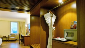 A hotel room with a wooden ceiling, a bed, a desk with a chair, a robe hanging on a hook, and a television on a stand. - Peerless Hotel Kolkata (Kolkata)