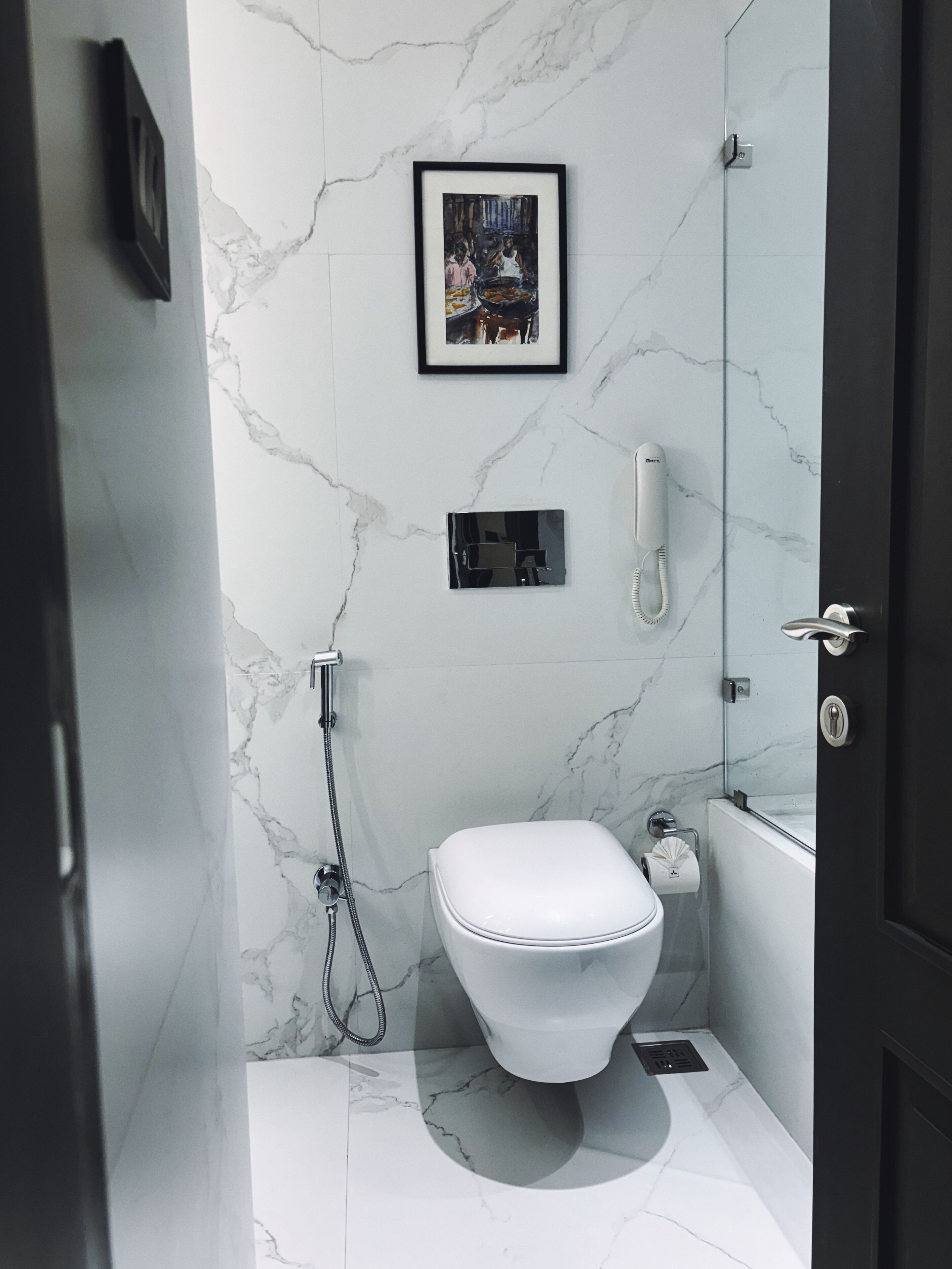 Une salle de bain moderne avec un sol en marbre, des toilettes blanches, un tableau encadré au mur et un téléphone fixé au mur.