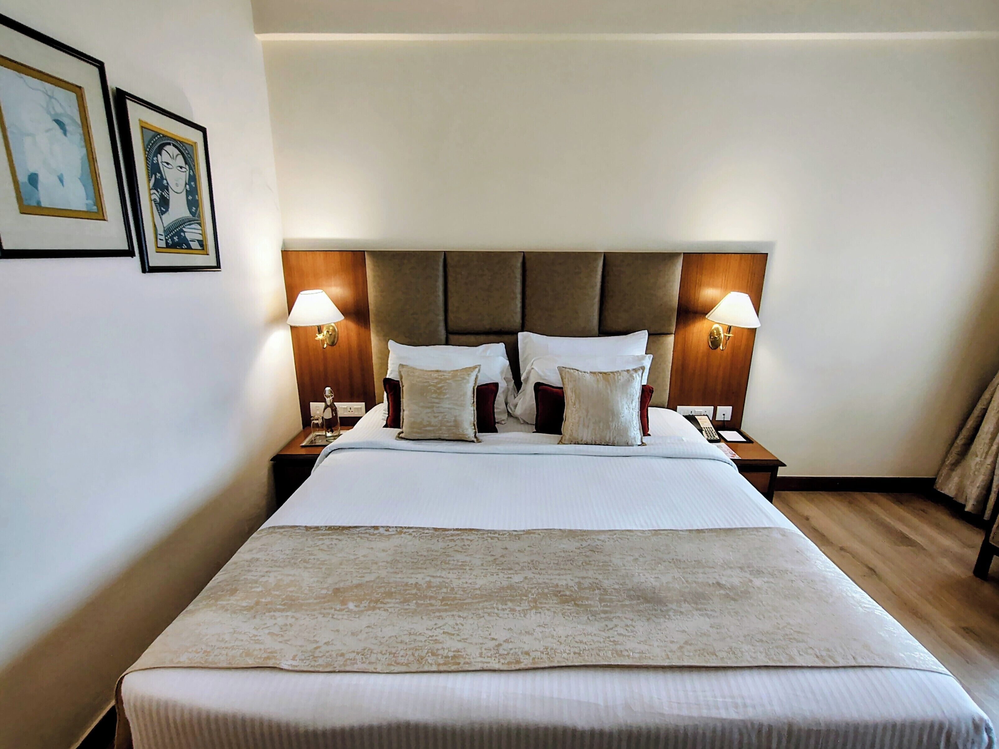 Quarto de hotel com uma cama grande, duas luminárias de cabeceira, uma cabeceira e duas fotos emolduradas na parede.