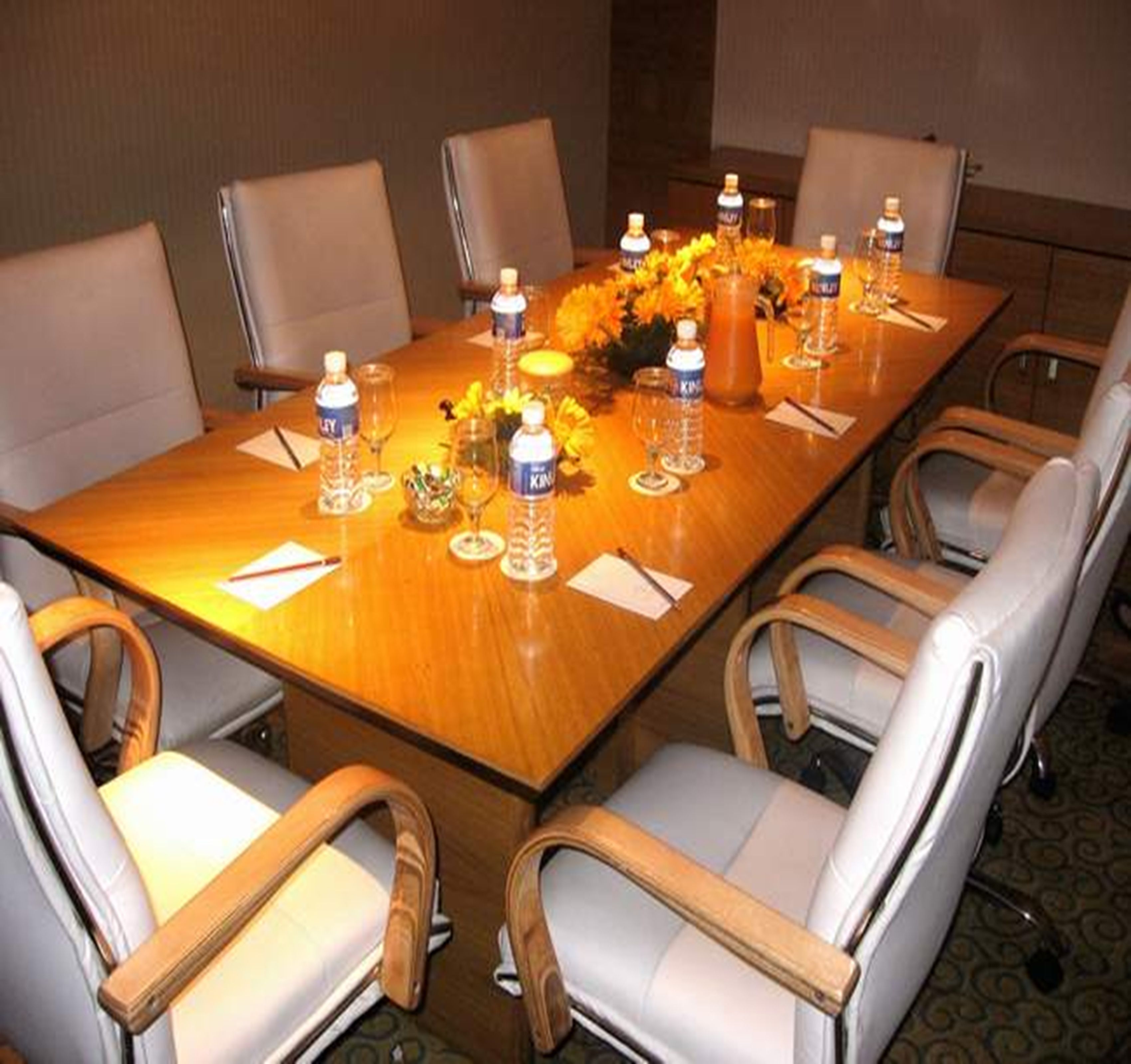 Un salón de conferencias con una larga mesa, seis sillas, botellas de agua, vasos y un jarrón con flores.