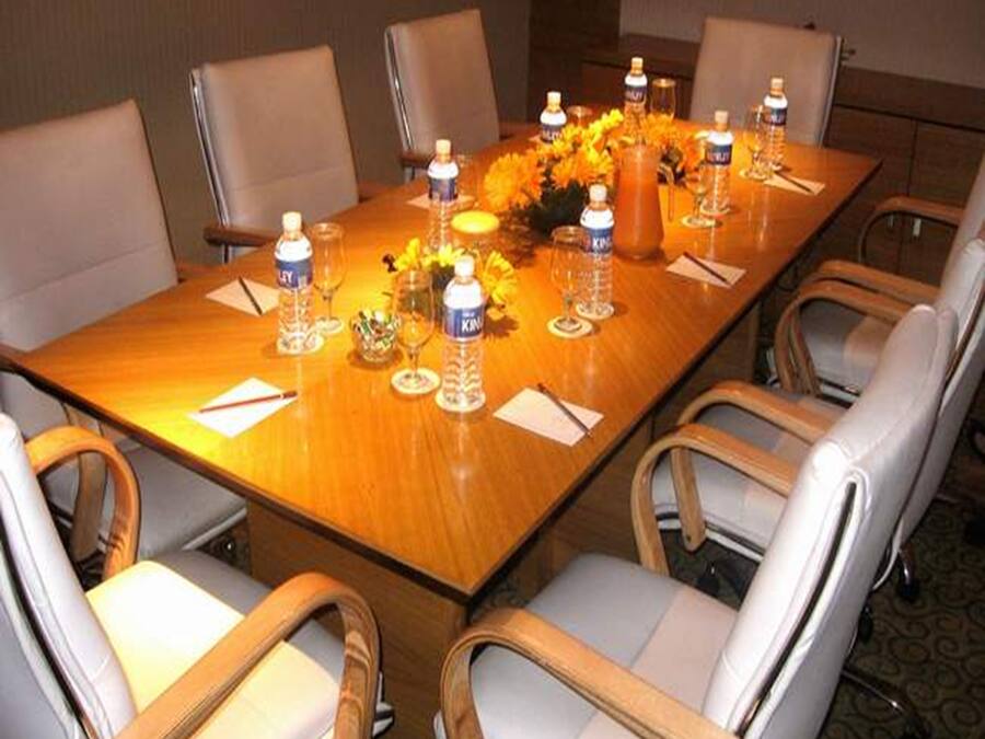 Un salón de conferencias con una larga mesa, seis sillas, botellas de agua, vasos y un jarrón con flores.