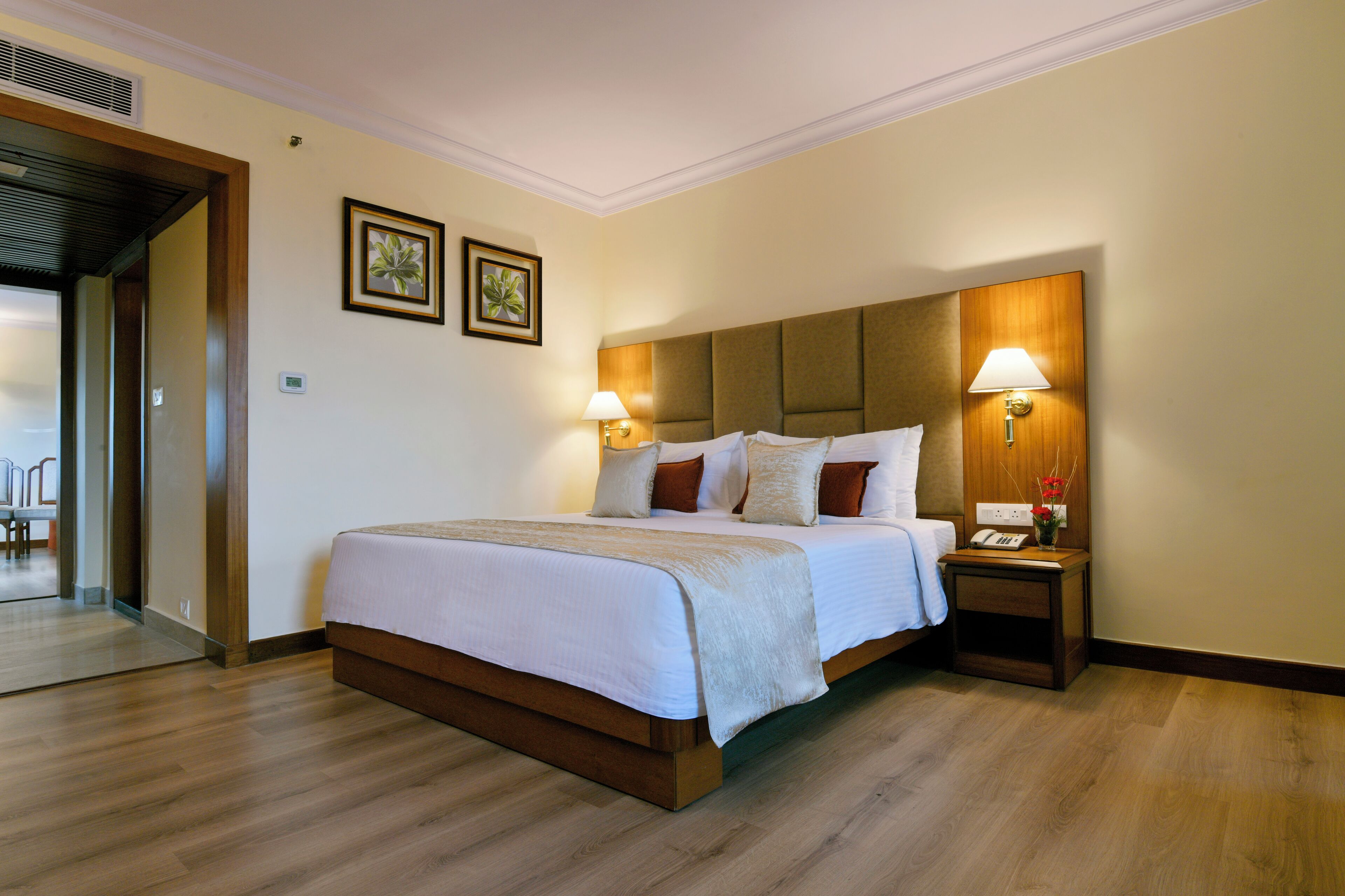 Quarto de hotel com cama grande, cabeceira de madeira, duas luminárias laterais, mesa de cabeceira com telefone e vaso, e porta que dá para um corredor.