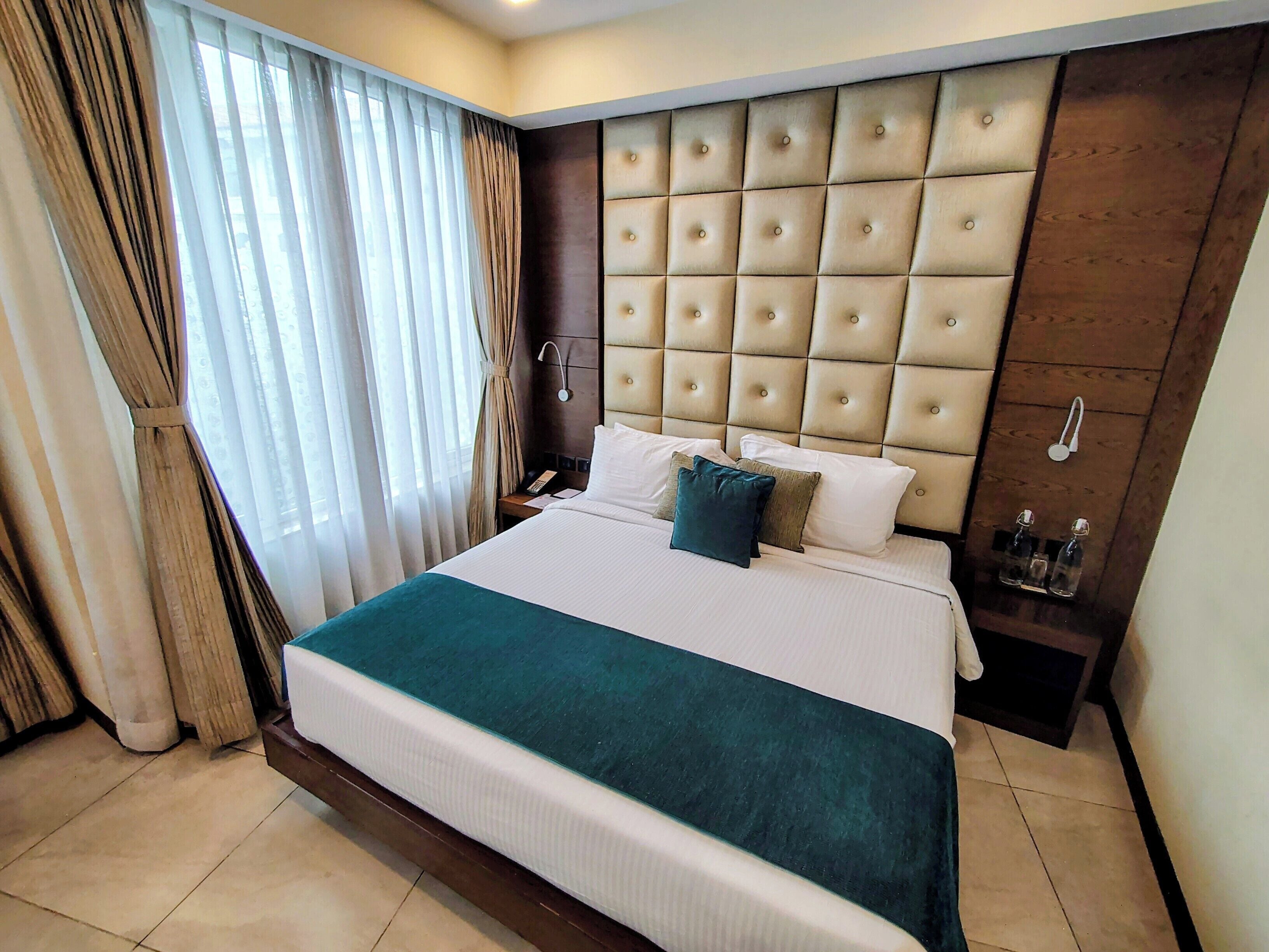 A hotel room with a large bed, a wooden headboard, a bedside table with a lamp, and a window with curtains.