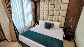 A hotel room with a large bed, a wooden headboard, a bedside table with a lamp, and a window with curtains.