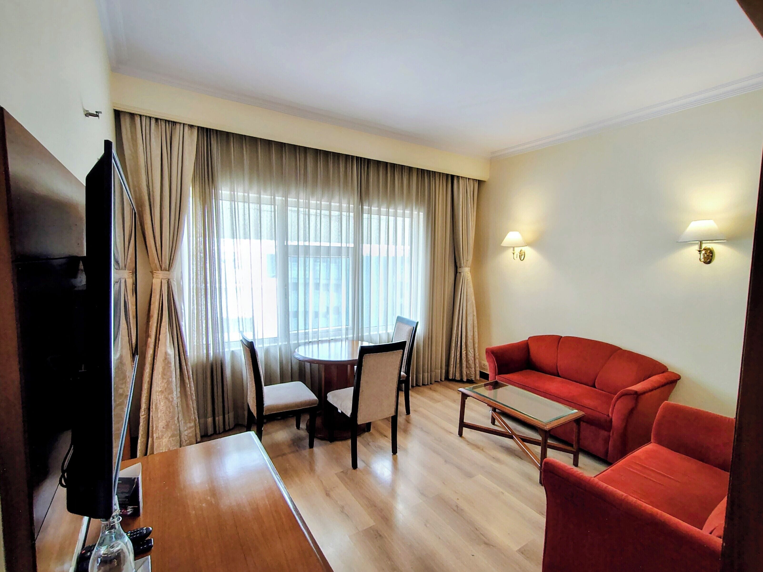 Une chambre d’hôtel avec une table à manger, des chaises, un canapé rouge, une télévision et des rideaux.