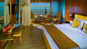 Quarto de hotel com cama, mesa de jantar com cadeiras, cesta de frutas e vista para o mar.