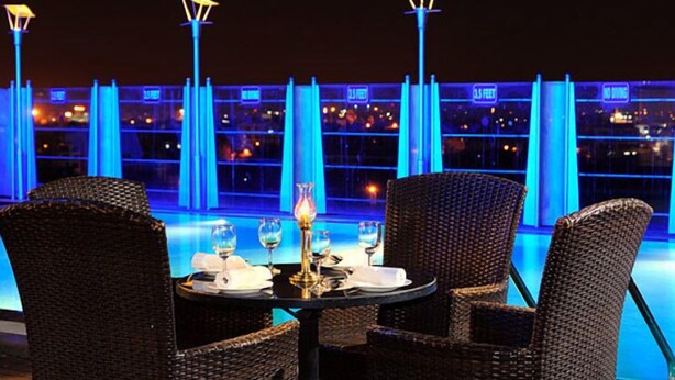 Un comedor con sillas de mimbre y una mesa dispuesta para dos personas, con vistas a una piscina iluminada en azul y un panorama urbano.