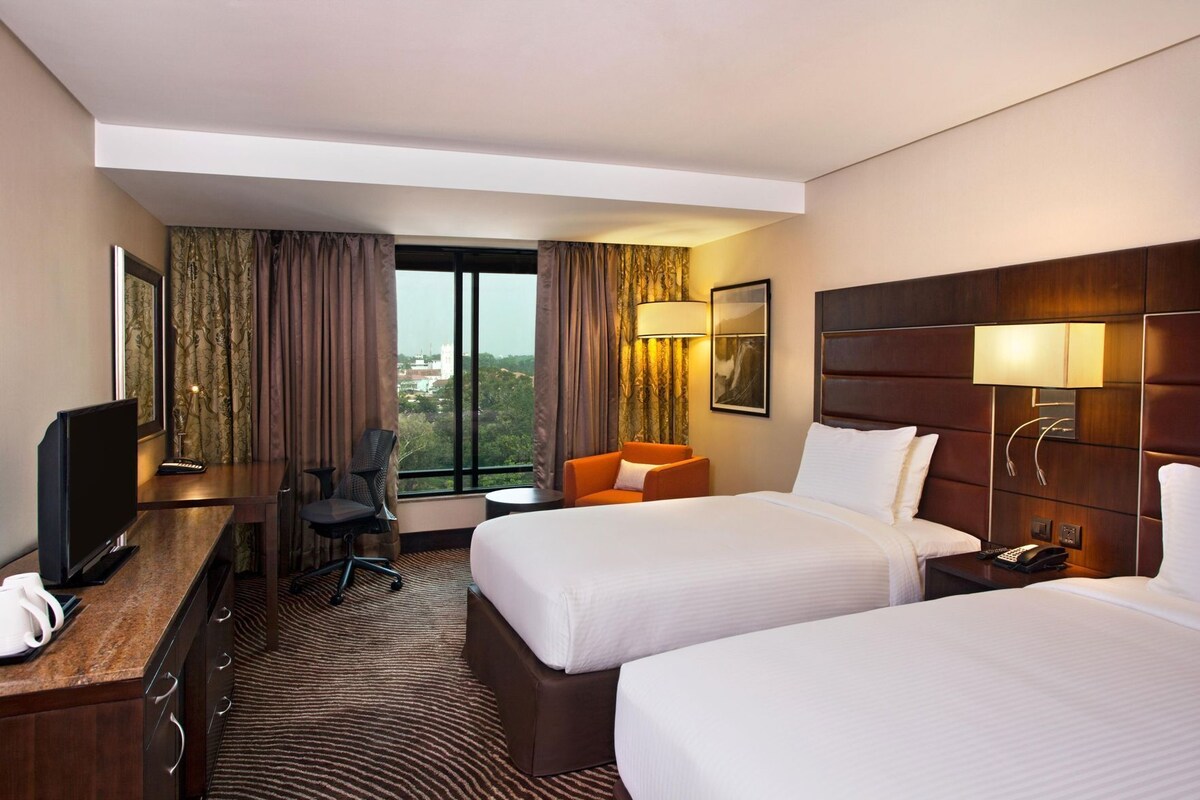 A hotel room with two beds, a desk with a TV, a chair, a small table, and a window with curtains.