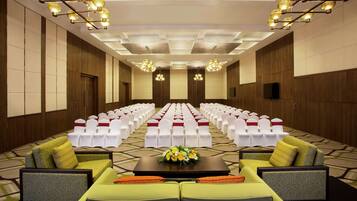 Un salón de conferencias con filas de sillas blancas, un escenario con un arreglo floral y un sofá con cojines.