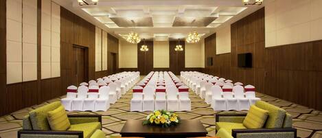 Un salón de conferencias con filas de sillas blancas, un escenario con un arreglo floral y un sofá con cojines.