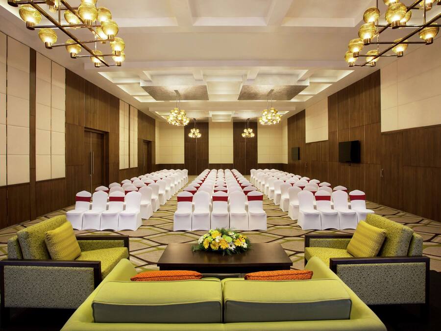Un salón de conferencias con filas de sillas blancas, un escenario con un arreglo floral y un sofá con cojines.