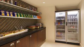 Una kitchenette con estantes llenos de aperitivos, salsas y bebidas, y un refrigerador con varias opciones.