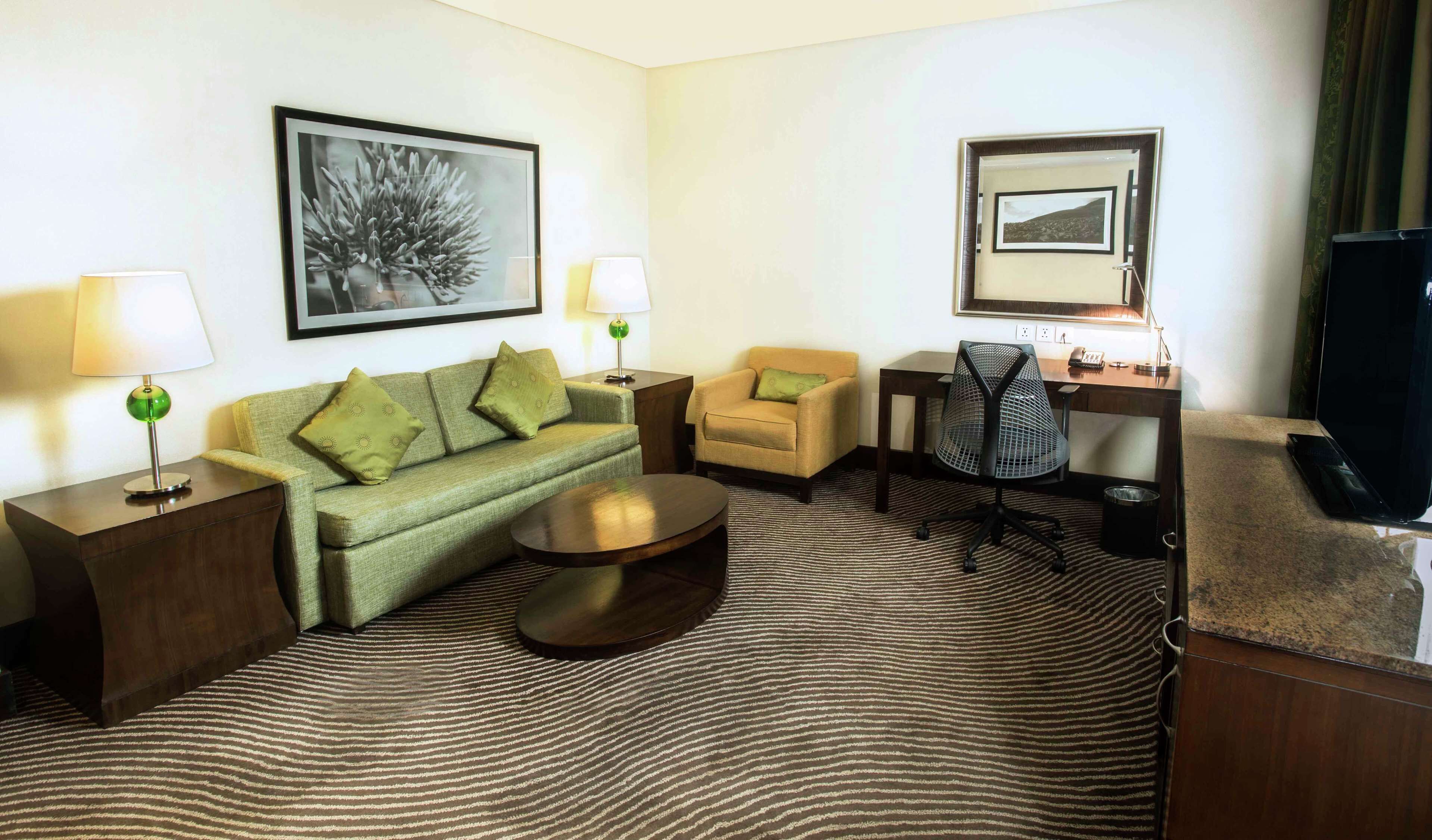 A hotel room with a green sofa, a round coffee table, a small armchair, a desk with a computer, a TV, and a zebra-patterned rug.