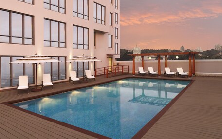 A rooftop pool with lounge chairs and umbrellas, surrounded by a building with large windows.