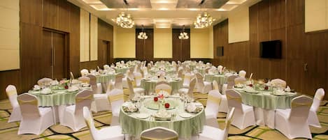 A banquet hall with multiple round tables, each set with white chairs and green tablecloths, featuring chandeliers and wooden paneling.