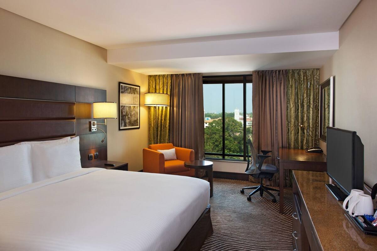 A hotel room with a large bed, a desk with a chair, a television, and a view of the city through the window.