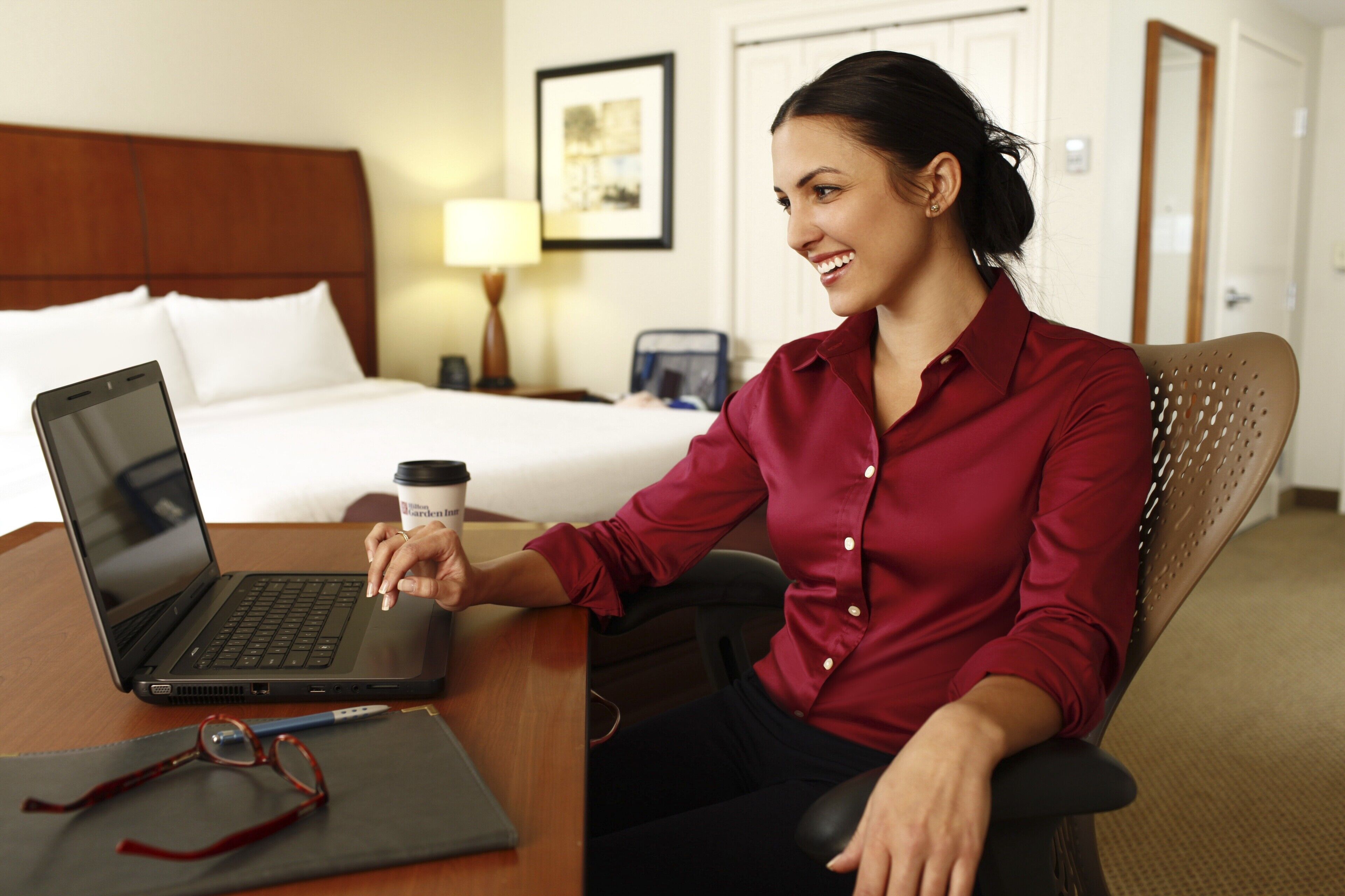 Une chambre d’hôtel avec un lit, un bureau avec un ordinateur portable, une tasse à café et une paire de lunettes.