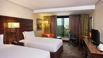 Quarto de hotel com duas camas, uma mesa de trabalho com televisão, uma cadeira, abajur e vista da cidade através de grandes janelas.