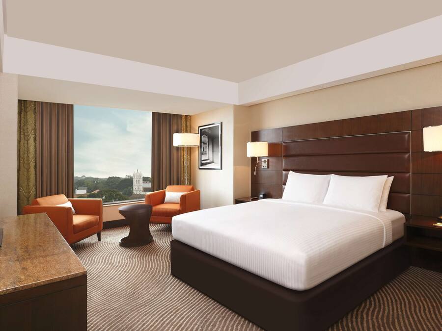 A hotel room with a large bed, two armchairs, a desk with a TV, and a view of the city.