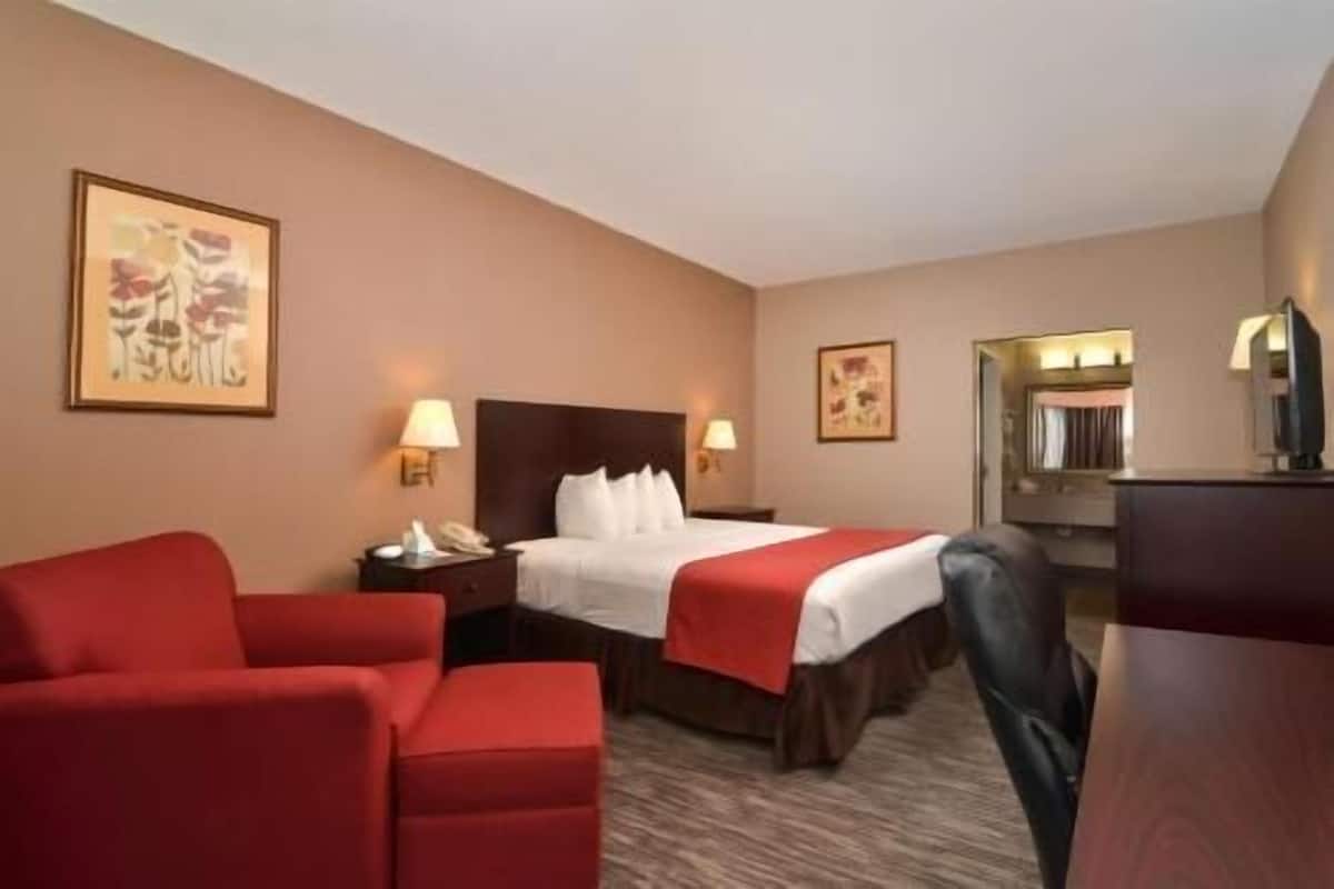 A hotel room with a bed, two bedside tables, a desk, a chair, and two red armchairs.