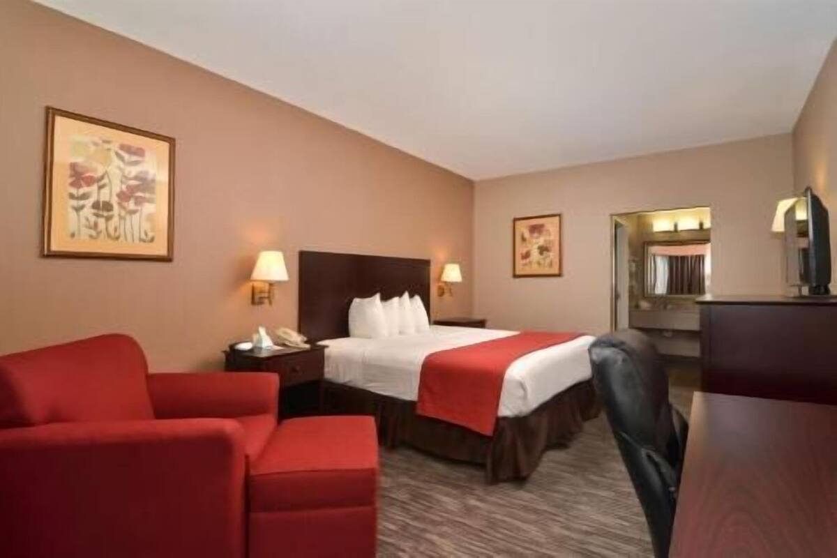A hotel room with a bed, two bedside tables, a desk, a chair, and two red armchairs.