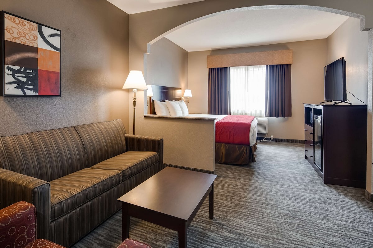 A hotel room with a sofa, a small table, a desk with a computer, a TV, and a bed with a red blanket.